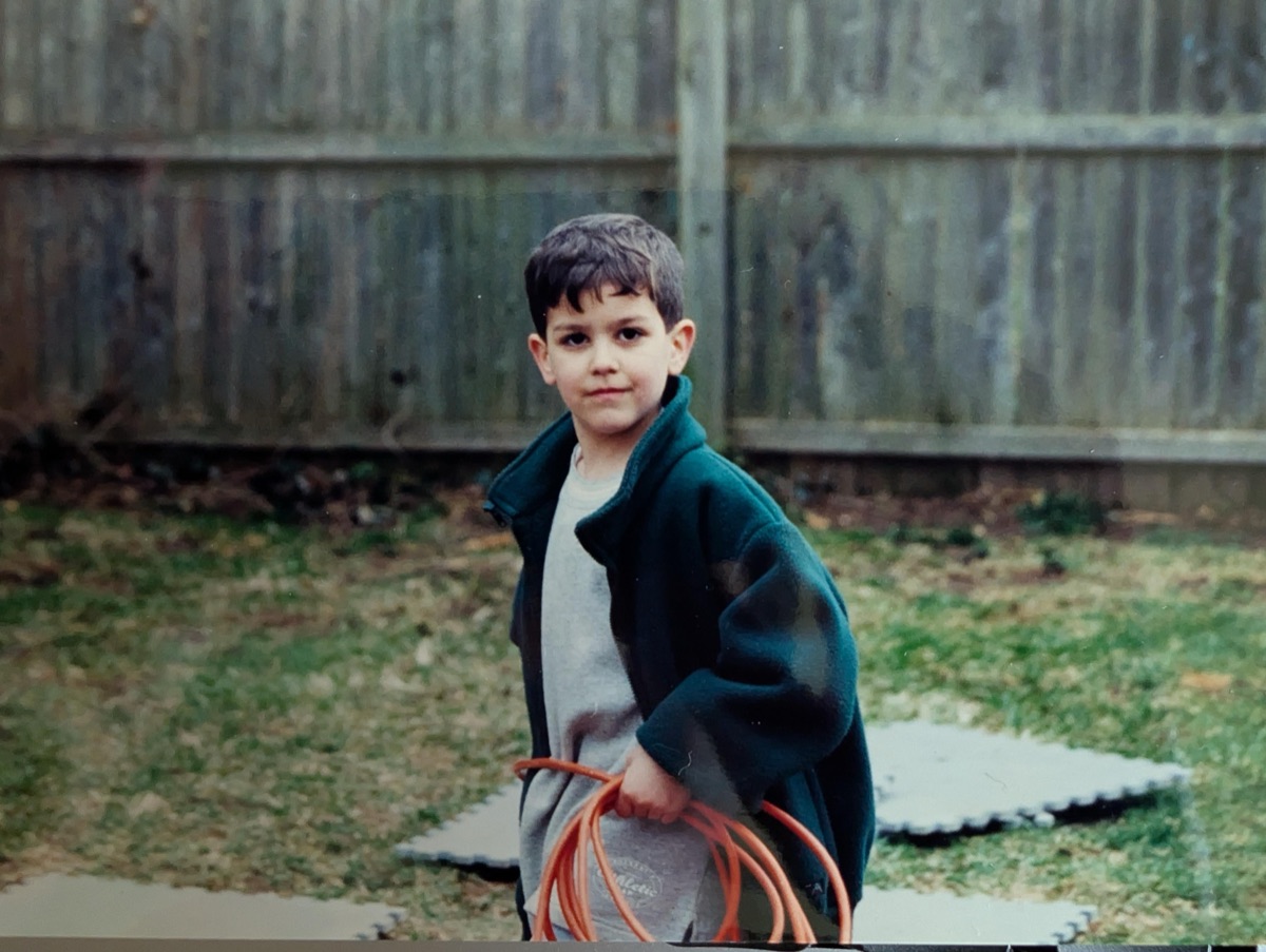 John as a young boy in a backyard, holding a coiled orange extension cord